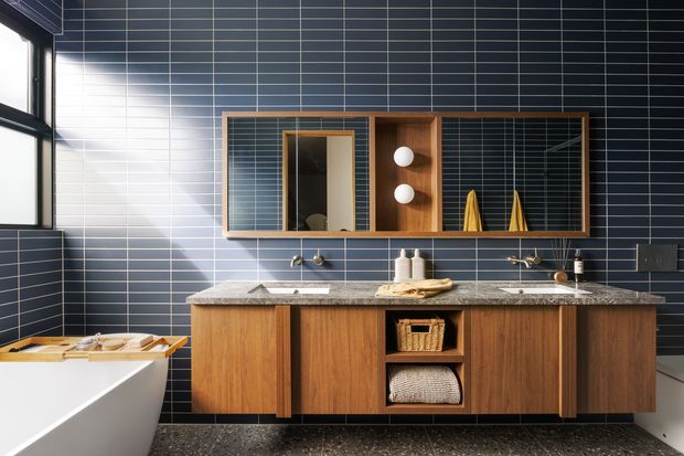 A stylish mid-century-inspired bathroom with a long double vanity in warm timber cabinetry, grey stone benchtop, twin basins and wall-mounted taps. The room features dark blue horizontal rectangular wall tiles, a large timber-framed mirrored cabinet with open shelving, integrated lighting, terrazzo-like dark flooring, and a partially visible freestanding white bathtub. Strong natural light enters from the window at left.