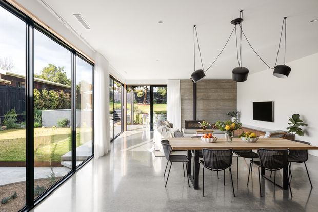 Bright open-plan dining and living area with floor-to-ceiling black-framed glazing along one side, polished concrete flooring, white walls and ceiling, a large timber dining table with black chairs, sculptural pendant lights, and a timber-toned feature wall around the television and fireplace. The glazing opens visually onto a landscaped garden.