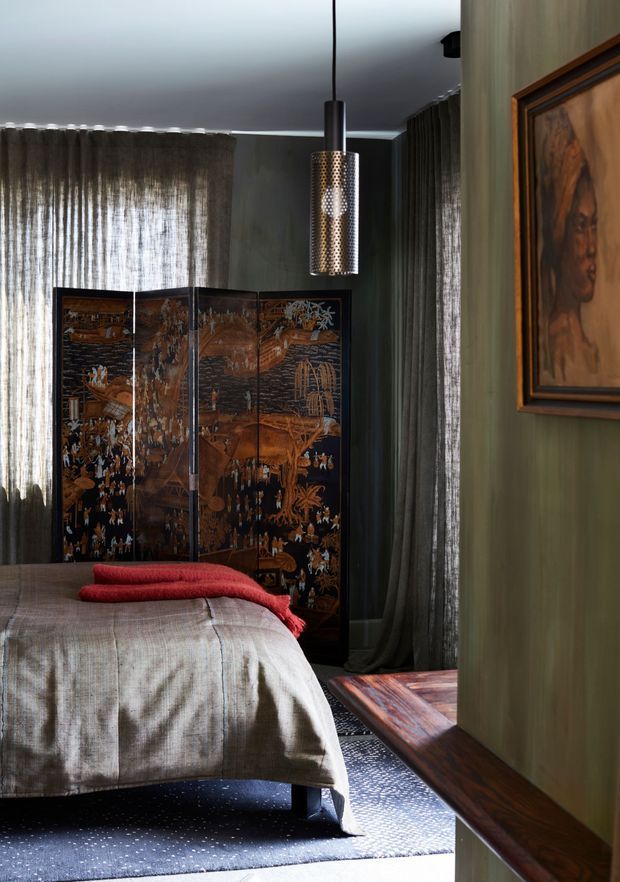 A moody, richly layered bedroom with a low upholstered bed, taupe-grey bedding, a folded red throw, dark patterned area rug, full-height sheer charcoal curtains, and deep green-grey wall panelling. A decorative dark wood folding screen stands behind the bed, while a perforated metal pendant light hangs centrally and framed artwork is visible on the right wall. The room has a refined, eclectic feel with warm timber detailing and dramatic low-key lighting.