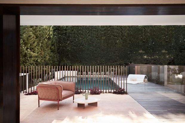 A refined outdoor terrace viewed through a large dark timber-framed opening, featuring a pale paved patio, blush upholstered lounge chair, small round coffee table, timber slatted privacy fencing, and a swimming pool beyond. A timber deck walkway extends alongside the pool, with dense clipped greenery forming a private backdrop.