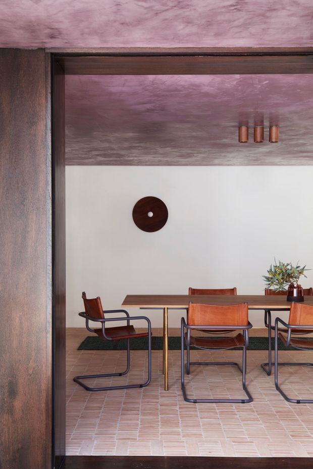 A refined dining room viewed through a dark timber-framed opening. The room features a long timber dining table, leather sling chairs with dark metal frames, pale brick or terracotta tile flooring, white walls, and a textured mauve-pink ceiling. A simple round dark wood wall piece and a small floral arrangement complete the understated setting.