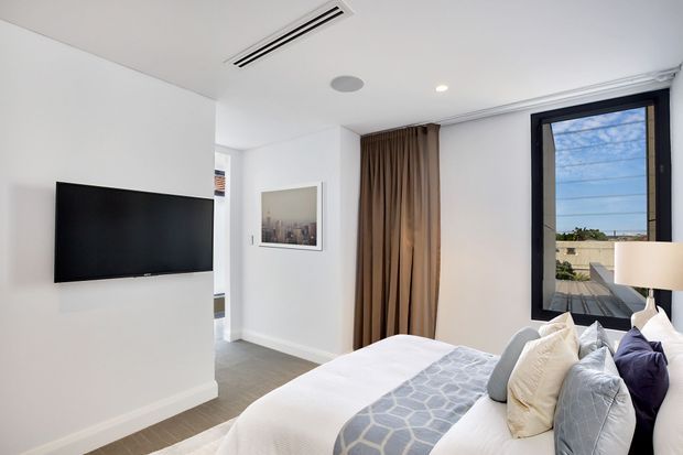 Bright contemporary bedroom with a large upholstered bed, layered neutral and blue-grey bedding, floor-to-ceiling brown curtains, white walls, grey carpet, recessed lighting and black-framed window glazing. A wall-mounted television and framed artwork are visible, with an urban rooftop outlook through the window.