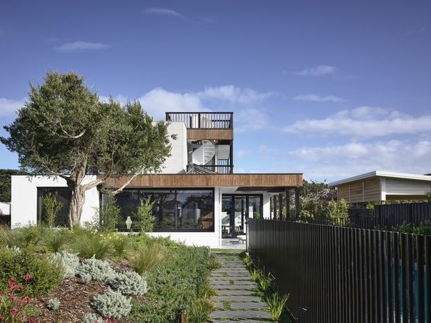 Contemporary multi-level residence viewed from a landscaped front garden, featuring a flat-roofed ground-floor pavilion, extensive dark-framed glazing, timber-look upper cladding, a rooftop terrace, and a sculptural external spiral stair. A paved stepping-stone path leads through dense coastal-style planting toward the entry, with a dark vertical-slat boundary fence in the foreground.