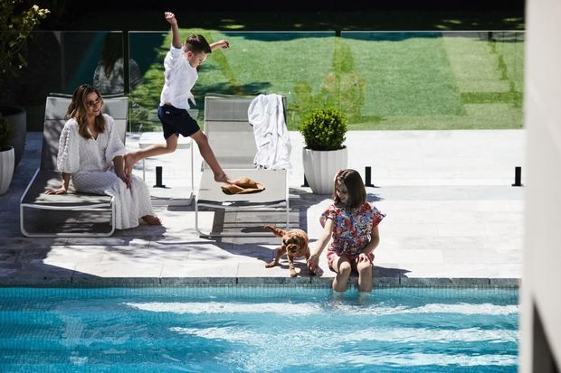 A bright outdoor pool area with a modern white-paved terrace, glass balustrade, streamlined loungers, potted planting, and children enjoying the pool with a dog. The scene has a clean contemporary resort-like feel and strong indoor-outdoor connection.