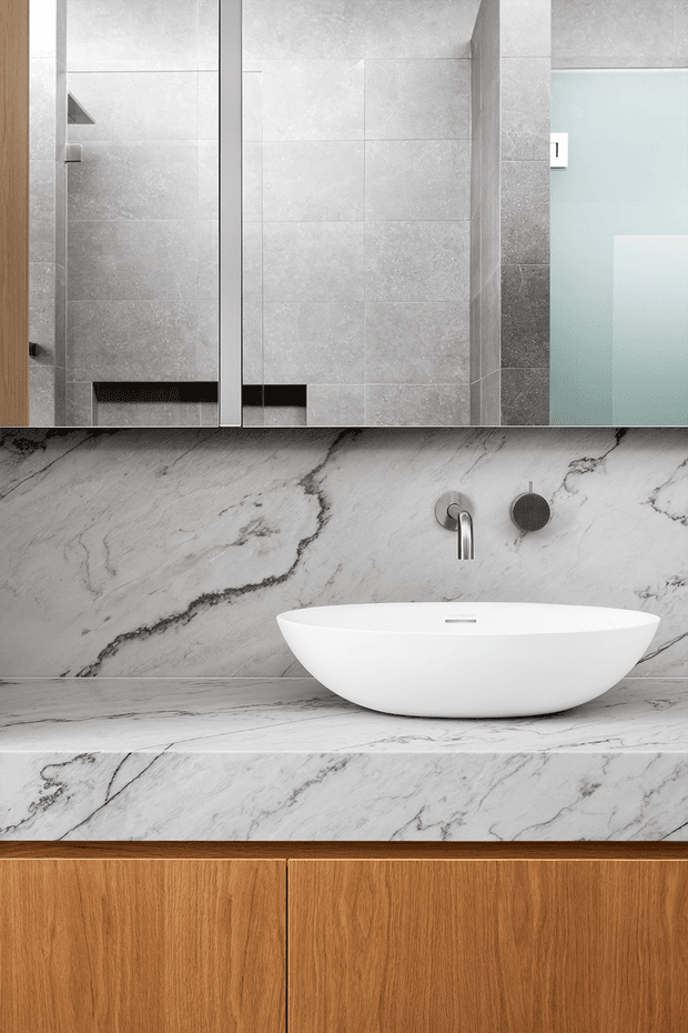 Close-up of a refined contemporary bathroom vanity. A white vessel basin sits on a dramatic grey-veined marble or stone benchtop and matching splashback, with wall-mounted chrome tapware and a dark circular control. Warm timber cabinetry forms the vanity base. Grey tiled shower walls, a frameless glass shower screen, and a mirrored cabinet are visible above.