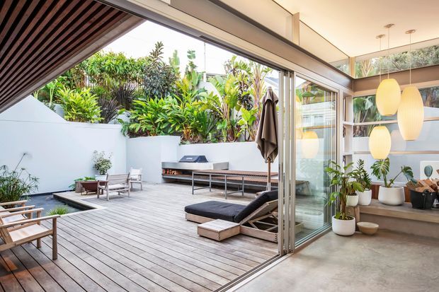 A contemporary indoor-outdoor living space opening onto a spacious timber deck and landscaped courtyard. Large retractable glass doors connect the polished concrete interior to an outdoor lounge with sun loungers, timber seating, a built-in barbecue, and lush tropical planting. A slatted timber ceiling and sculptural pendant lights add warmth and texture.
