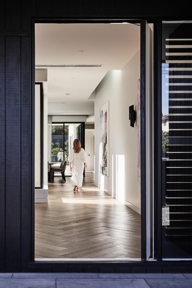 View through a dark-framed exterior doorway into a bright, refined residential hallway. The interior features pale walls, recessed ceiling lighting, large artwork, a herringbone timber floor, and full-height glazing at the far end. A woman is walking toward an adjoining living area, while exterior black cladding and a partially visible dark louvred screen frame the entrance.