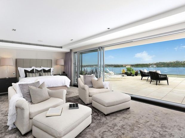 A bright, luxurious bedroom suite with a large upholstered bed, matching armchairs and ottoman, and a broad area rug. Full-height sliding glass doors open to a spacious waterfront terrace with outdoor seating and panoramic water views. The interior features a neutral palette, streamlined built-in window framing, and a low contemporary ceiling.