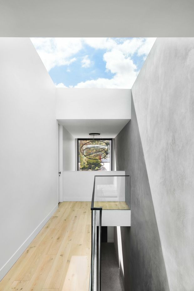A minimalist interior landing or hallway with pale timber flooring, white plaster walls, a textured grey feature wall, and a black-framed glass balustrade overlooking a stairwell. A large open skylight brings in daylight and views of blue sky, while a window at the end of the corridor overlooks trees and contains a sculptural circular pendant light.