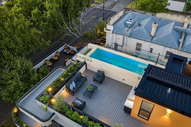 Elevated aerial view of a rooftop terrace with a long rectangular swimming pool, grey decking, outdoor lounge seating, planter beds, and warm accent lighting. The terrace is integrated with a contemporary dark-clad rooftop structure and overlooks a leafy urban street and neighbouring building roofs.