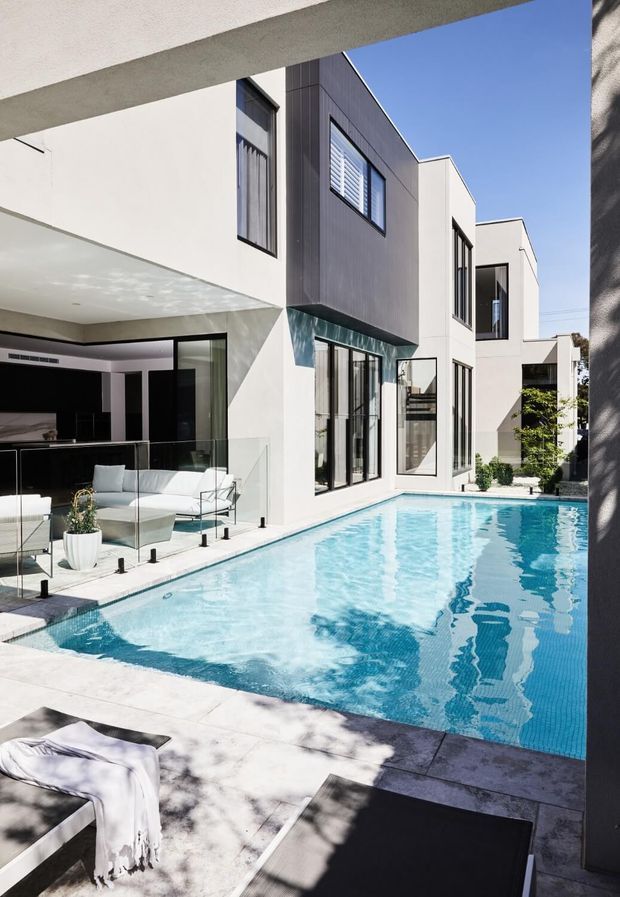 A bright contemporary multi-storey home arranged around a narrow rectangular swimming pool. The courtyard-facing facade uses crisp white rendered walls, dark vertical cladding, extensive black-framed glazing and projecting box forms. A covered outdoor lounge with glass balustrading sits beside the pool, while pale stone paving and strong sunlight create a clean, resort-like atmosphere.