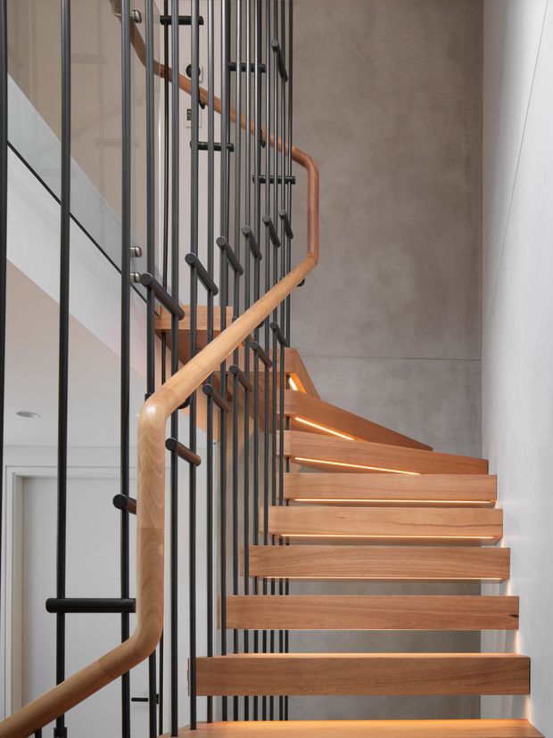 Interior architectural photograph focused on a contemporary open-riser staircase. Warm timber treads and a rounded timber handrail contrast with slender black metal vertical balusters, set against a smooth grey concrete wall. Integrated linear lighting glows beneath several stair treads, and a glazed balustrade is visible at the side. The composition emphasises the stair detailing and vertical circulation element.
