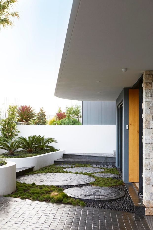 Contemporary residential entry courtyard viewed from outside, featuring a broad white soffit, dark-framed glazing, a warm timber pivot door, and a textured stacked-stone wall. Curved raised garden beds contain sculptural cycads and other planting, while circular stepping-stone pads and dark pebble landscaping lead toward the entrance.