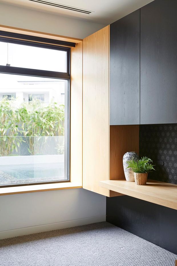 A compact, contemporary built-in study nook or home office with a large black-framed window, warm timber wall lining, dark textured cabinetry, and a floating timber desk. A patterned black feature backsplash, decorative vase, and small plant add detail, while grey carpet and a neutral ceiling create a calm, refined interior.