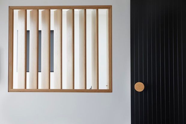 Minimal interior architectural detail featuring a rectangular timber-framed internal window or screen with evenly spaced vertical timber slats. A black ribbed door or panel with a round timber knob is visible at right, set against a pale grey wall. The image emphasises clean lines, natural timber and contrasting dark joinery.