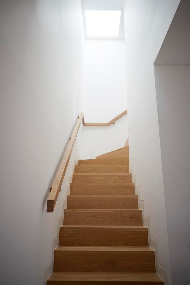A minimalist interior stairway with warm timber stair treads and matching timber handrails enclosed by crisp white walls. A rectangular skylight provides soft natural light from above, emphasizing the narrow vertical stairwell.