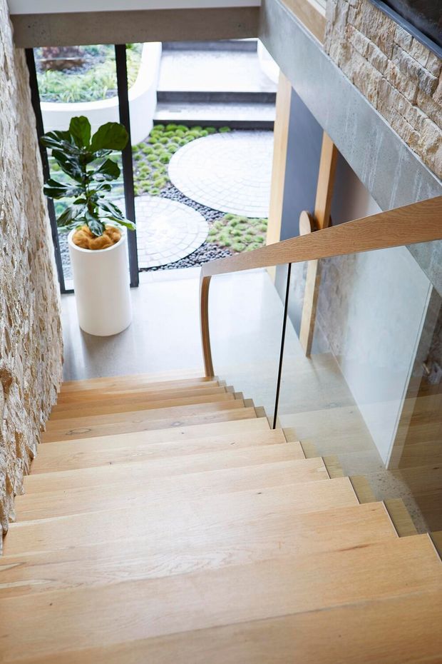 A bright, contemporary stairway descends through a narrow double-height entry space. The stairs are finished in pale timber, with a slim timber handrail and clear glass balustrade. Rough-textured stacked-stone walls contrast with smooth light walls and flooring. A tall potted plant sits at the landing, overlooking a landscaped courtyard with circular pale paving and pebble planting.