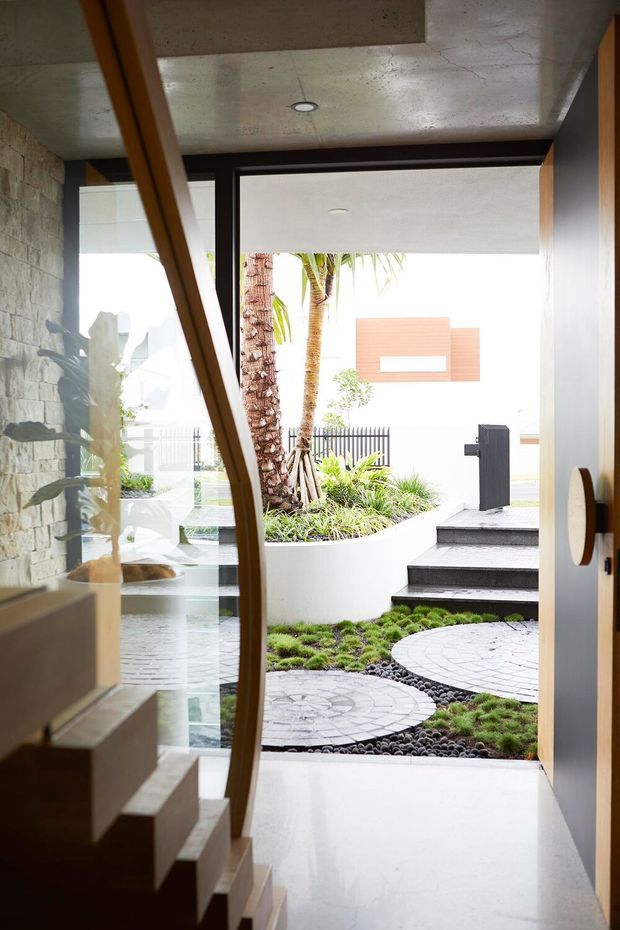 View from a contemporary interior entry toward a landscaped courtyard, with a sculptural curved timber stair and glass balustrade at left, exposed concrete ceiling, stone feature wall, polished light floor, and dark-framed glazing. The courtyard features curved white planters, tropical palms, pebble landscaping, circular patterned paving, and steps leading outside.