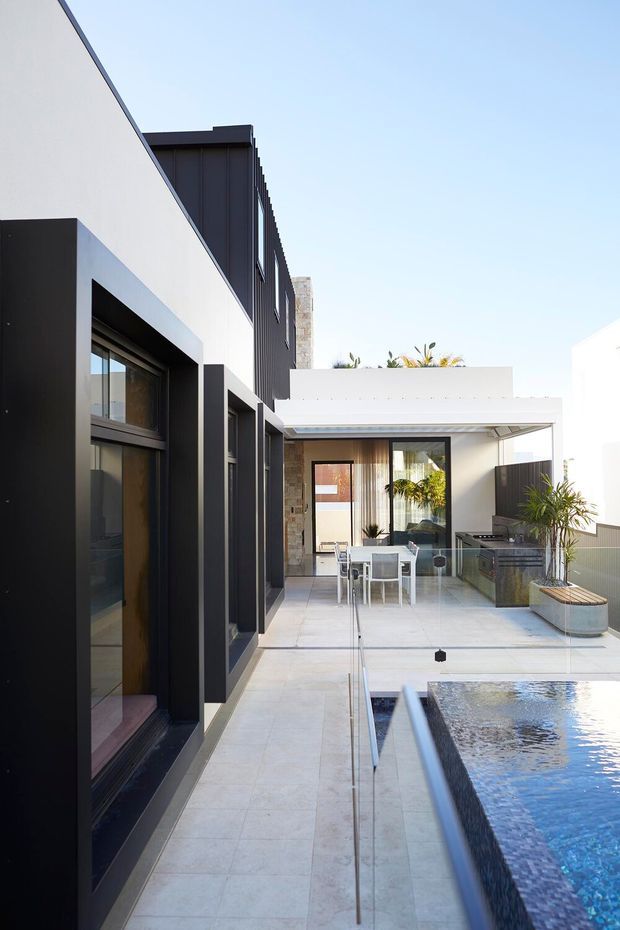 A contemporary courtyard terrace viewed alongside a modern house, with strong black-framed glazing, white rendered walls, dark vertical cladding, pale stone paving, and a glass-edged swimming pool. An outdoor dining area sits beneath a flat-roofed covered terrace, with potted palms adding greenery.