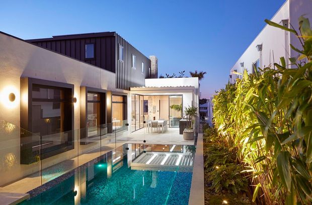 Contemporary multi-level residence arranged around a narrow turquoise swimming pool, with crisp white rendered walls, dark vertical metal cladding, extensive glazing, a glass pool balustrade, and lush screening planting along one side. The image is taken from an exterior poolside terrace at dusk, showing a bright open living or dining area beyond the pool.
