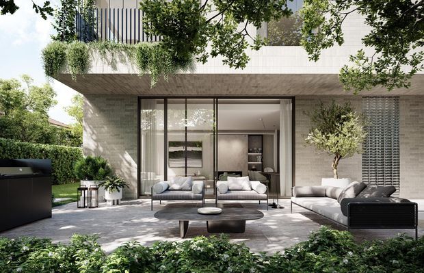 A contemporary residential courtyard viewed from the garden, featuring a pale textured masonry facade, expansive dark-framed glazed sliding doors, and a covered outdoor lounge with sofas, coffee tables, and potted planting. Dense greenery surrounds the paved terrace, while a black built-in outdoor kitchen or barbecue sits to the left. The upper level includes vertical screening and cascading plants, creating a refined indoor-outdoor connection.