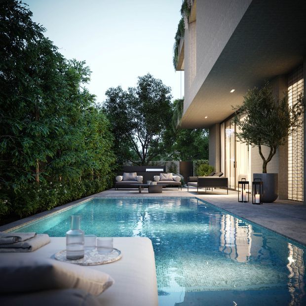 A contemporary outdoor pool courtyard beside a modern multi-level residence. The image shows a long rectangular swimming pool with pale blue mosaic-like finish, a paved pool deck, outdoor lounge seating, a low table, lanterns, dense landscaped screening, and a covered cantilevered upper floor with recessed lighting. Floor-to-ceiling glazing and a vertical textured screen define the house edge, while a cushioned daybed or sun lounger occupies the foreground.