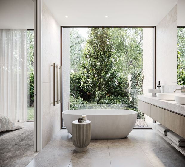 Bright, contemporary bathroom with a freestanding oval bathtub positioned in front of expansive floor-to-ceiling glazing overlooking a leafy garden. A long floating timber-toned vanity with an integrated pale stone benchtop and vessel basins lines the right wall. Large pale floor and wall tiles, minimal recessed lighting, brushed metal towel rails, soft grey textiles, and a small ribbed stool create a calm, refined atmosphere. A bedroom is partially visible through the open connection at left.
