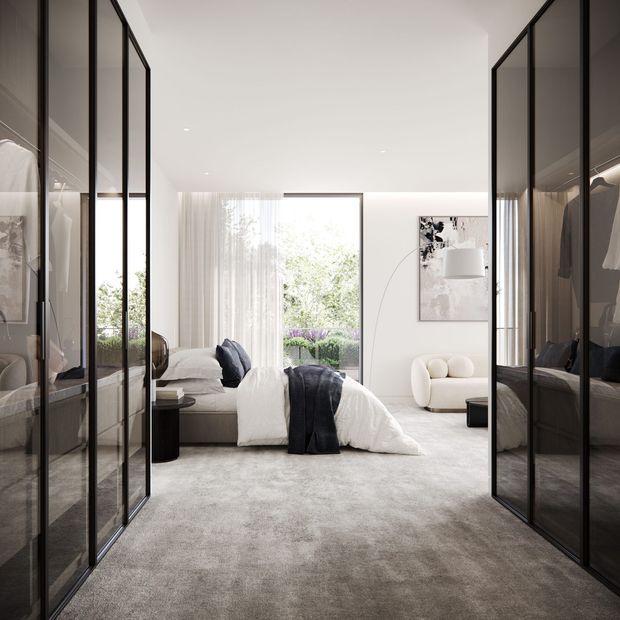 A bright, refined contemporary bedroom viewed through open, floor-to-ceiling glass-front wardrobes. The room features a low upholstered bed with white bedding and dark charcoal throws, pale grey carpet, sheer curtains, a large window overlooking greenery, a sculptural white lounge chair, and minimalist artwork with a floor lamp. Black-framed wardrobe glazing creates strong vertical lines on both sides of the room.