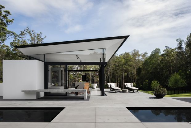 Contemporary single-storey residence with a dramatic flat cantilevered roof, extensive black-framed glazing, and a white minimalist facade. A broad light-grey paved terrace surrounds a dark reflecting pool, with outdoor lounge furniture positioned beside a wooded landscape.