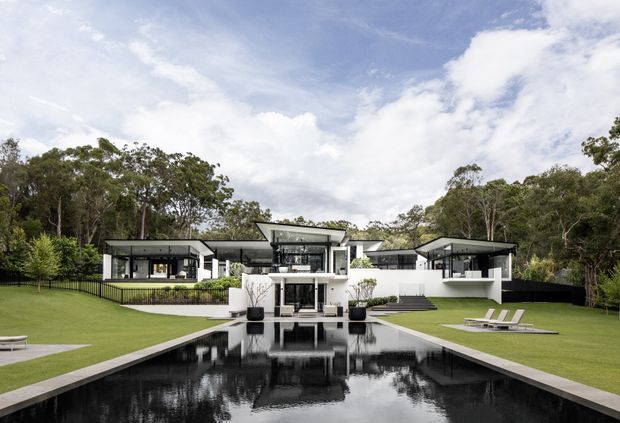 A large contemporary residential property viewed from the rear across an expansive rectangular reflecting pool. The white and dark-framed house has a low, horizontally composed form with several projecting flat-roof wings, extensive glazing, terraces, and a central upper-level feature volume. Manicured lawns, outdoor seating, planted beds, and mature trees surround the building.
