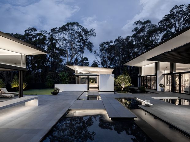 A dramatic twilight view of a contemporary multi-wing residence arranged around a series of long reflective pools and paved courtyards. Clean white geometric volumes, extensive dark-framed glazing, deep roof overhangs, and a prominent black slatted canopy define the architecture. Subtle integrated lighting illuminates the pale stone paving, water features, and landscaped edges, with dense mature trees forming a dark backdrop.