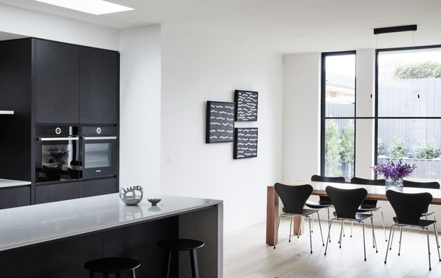 Bright contemporary open-plan kitchen and dining area with matte black full-height cabinetry, integrated appliances, a large pale stone island, and black stools. A timber dining table with black chairs sits beside tall black-framed windows, while graphic wall art and a linear pendant add restrained decorative accents.