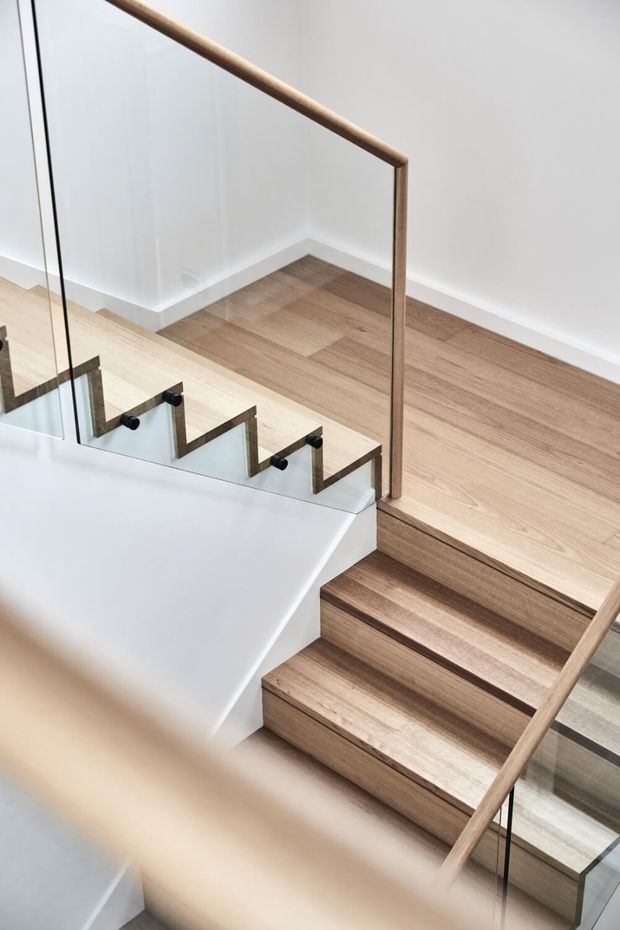 A contemporary stairway with light timber treads and flooring, clear glass balustrades, and slim timber handrails. White walls and black metal balustrade fittings create a clean, minimalist composition viewed from above.