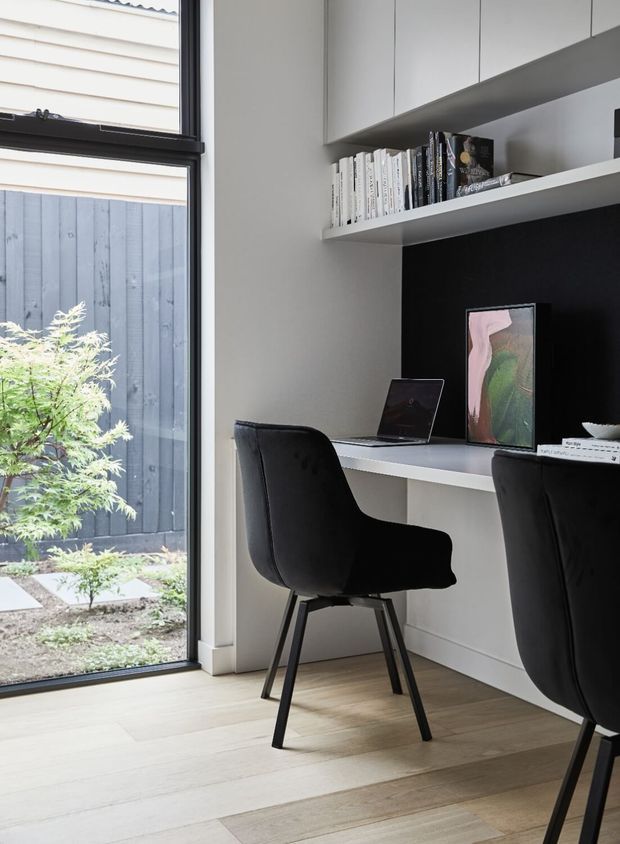 A compact, contemporary home office or study nook with a built-in white desk, open shelving, overhead cabinetry, books, artwork and two black upholstered chairs. Full-height glazing opens to a small landscaped courtyard, while pale timber flooring and a dark feature backing create contrast.