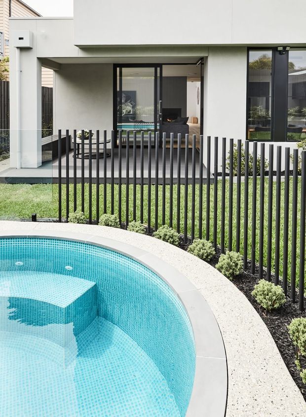 A contemporary residential backyard featuring a curved swimming pool with turquoise mosaic tiling and integrated bench steps in the foreground. A slim black vertical-bar fence separates the pool from a manicured lawn and a modern covered outdoor entertaining area with dark-framed glazing, an outdoor fireplace, seating, and a dining table. The house has a clean white rendered facade with broad, minimalist openings and a flat roofline.