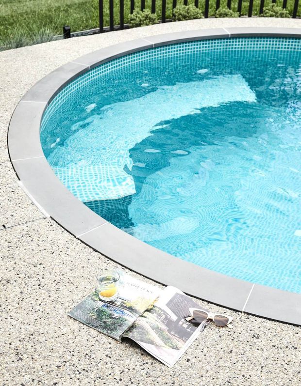 Close-up of a curved swimming pool with bright turquoise water, small blue mosaic tile lining, and a smooth light-grey coping edge. The pool is surrounded by a pale exposed-aggregate concrete deck, with a black metal fence and landscaping beyond. An open magazine, sunglasses, and a drink sit on the deck in the foreground.