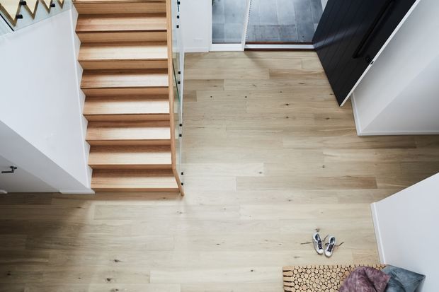 Overhead view of a contemporary residential stairway and landing. Light natural timber flooring continues beneath open-riser timber stairs, with a clear glass balustrade, white walls, and a dark-framed doorway leading toward a grey tiled area. A small timber log-style mat and footwear sit near the entry.
