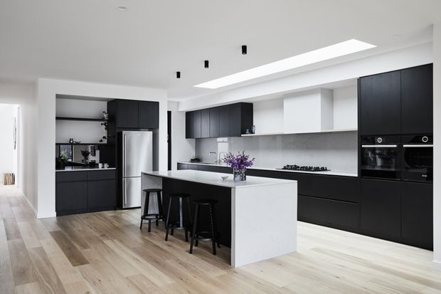 Bright contemporary kitchen with matte black full-height cabinetry, integrated black appliances, a large white island with waterfall-style sides, pale timber flooring, and a long linear skylight or ceiling recess. Three black stools sit at the island, with open shelving and a stainless-steel refrigerator visible at the rear.