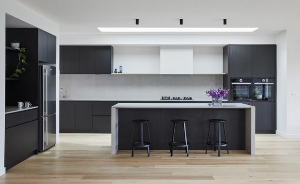 A contemporary open-plan kitchen with a large dark island and three black stools, floor-to-ceiling dark cabinetry, integrated stainless-steel refrigerator, built-in ovens, a pale stone-look splashback and benchtop, and light timber flooring. White walls and a recessed linear ceiling light create a clean, high-contrast interior.