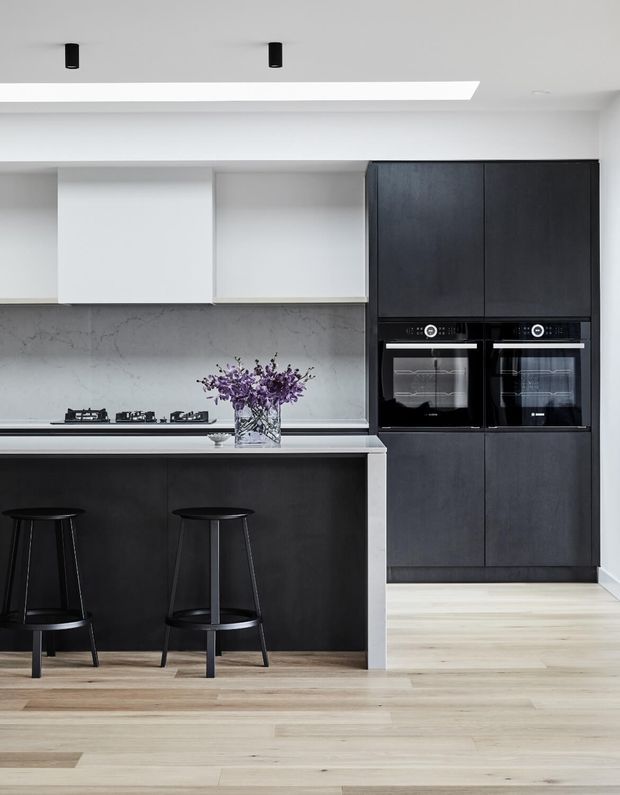 A contemporary kitchen with a large dark charcoal island, integrated black cabinetry and twin built-in ovens. The kitchen features a pale veined stone backsplash and benchtop, white overhead cabinetry, light timber flooring, black bar stools, and a long recessed ceiling light.