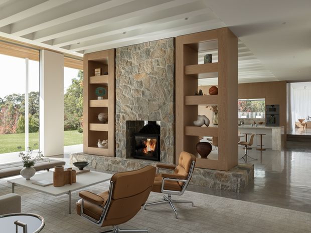 A spacious open-plan living room centred on a substantial natural stone fireplace with integrated warm timber display shelving. Mid-century style leather lounge chairs and a low coffee table sit on a large rug, while full-height glazing opens views to a lawn and trees. The room extends into a bright contemporary kitchen and dining area, with exposed white ceiling beams and polished concrete flooring creating a refined indoor-outdoor feel.