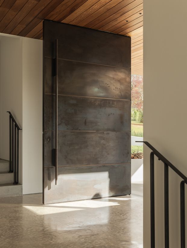 Interior view of a refined entryway featuring a tall, pivoting dark patinated metal door with horizontal panel divisions. The door is framed by light plaster walls, black metal stair railings, a warm timber-lined ceiling, and a polished concrete floor. Daylight enters from the open doorway, with a landscaped exterior visible beyond.