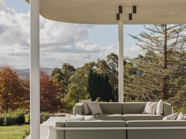 A covered outdoor terrace with a large built-in or modular sectional sofa overlooking a landscaped garden and distant wooded hills. The terrace has a flat concrete canopy supported by slender white columns, with simple dark cylindrical ceiling lights. Neutral upholstery and pale architectural surfaces create a calm, contemporary outdoor setting.