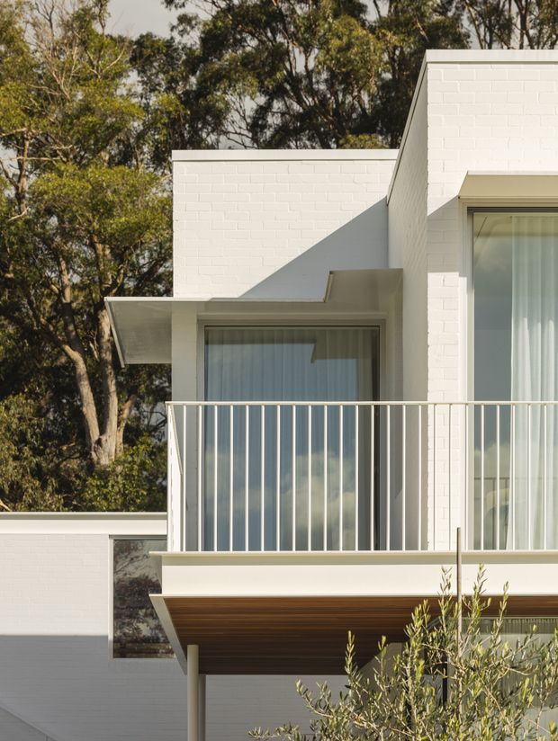 Close-up exterior view of a contemporary white-painted brick house surrounded by mature trees. The facade features crisp geometric volumes, a low-pitched or flat roof form, large glazed doors and windows with sheer curtains, a projecting balcony with slender white metal balustrade, and a warm timber-lined soffit beneath the balcony.