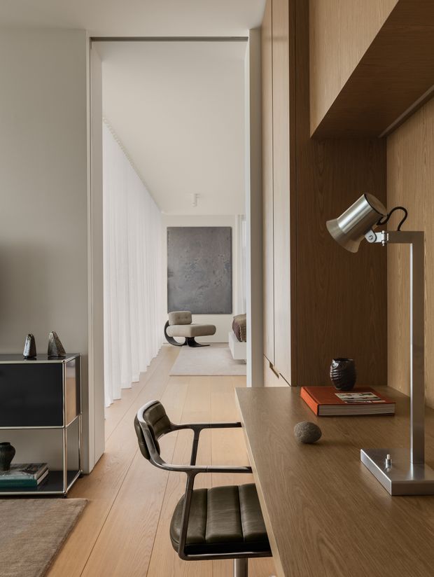A refined contemporary interior showing a compact built-in home office or study integrated into a timber wall unit. The foreground features a long wood desk, shelving, a silver task lamp, books and small decorative objects, paired with a dark leather office chair. Beyond the desk is a light-filled hallway or living area with pale timber flooring, white sheer curtains, a sculptural lounge chair and neutral artwork. The palette is warm and restrained, with clean lines and carefully integrated joinery.