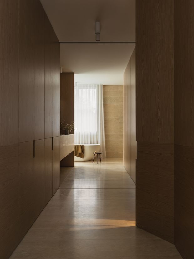 A softly lit minimalist interior hallway lined with full-height dark timber cabinetry and wall panelling. The corridor opens toward a bright sitting area with sheer curtains, a small stool, and warm beige stone or plaster wall surfaces. A smooth pale floor reflects natural light, creating a calm, refined atmosphere.