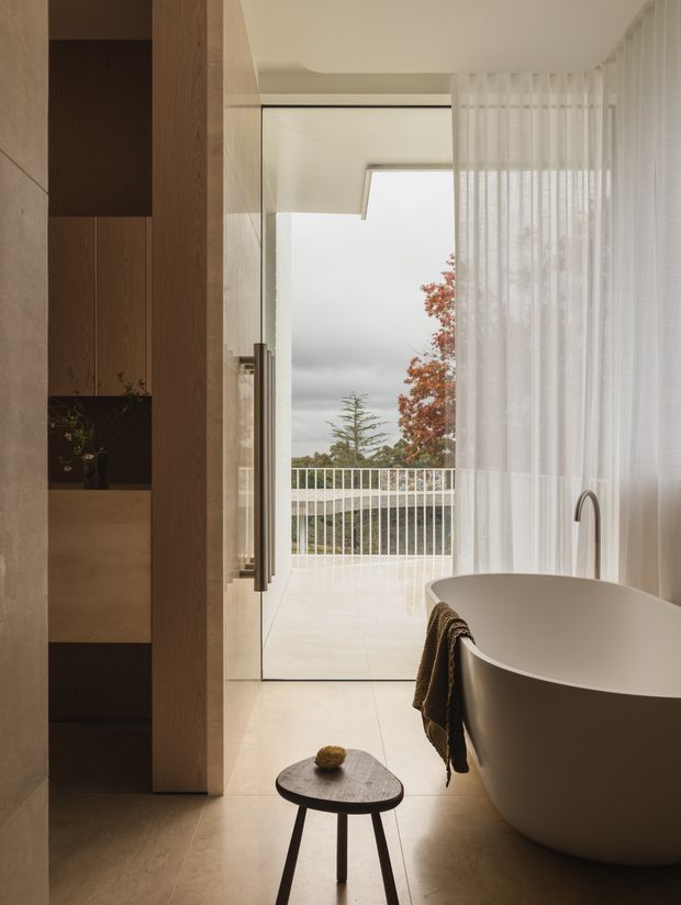 A refined contemporary bathroom with a freestanding oval bathtub positioned beside full-height sheer curtains and an open glass door to a balcony. Warm beige stone-look flooring and wall finishes are paired with dark timber built-in shelving, a slim black-edged glass door, and a minimalist floor stool. A sculptural curved faucet rises beside the bath, while soft natural light and muted outdoor foliage create a calm, luxurious atmosphere.