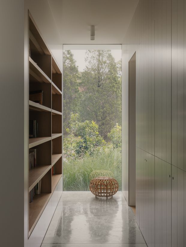 A narrow, refined interior passage or gallery with full-height glazing at the end opening onto dense garden planting. One side features extensive dark timber built-in shelving, while the opposite wall has tall, pale flush-panel cabinetry. Large polished light-grey stone or concrete floor slabs reflect the soft natural light. The restrained palette, clean lines, integrated joinery and indoor-outdoor connection create a calm, contemporary atmosphere.