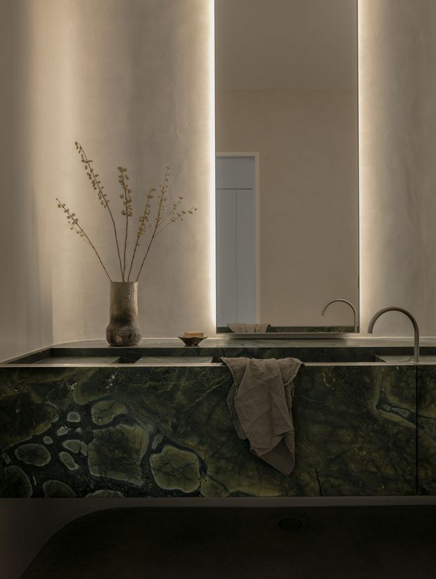 Moody, refined bathroom vanity viewed straight on, featuring a large backlit mirror, sculptural curved faucet, integrated dark green stone basin and cabinetry with dramatic natural veining, and warm softly textured plaster walls. A simple ceramic vase with dried branches and a draped neutral towel add understated organic detail.