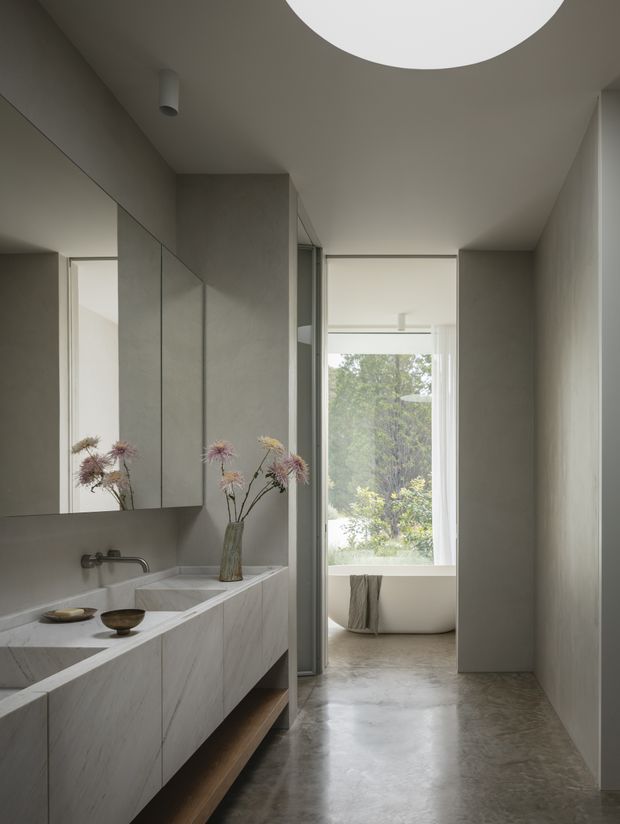 A refined minimalist bathroom vanity area opening directly toward a freestanding bathtub by a large window. The room features a long pale stone vanity with integrated basins, wall-mounted fixtures, full-height mirrored cabinetry, smooth grey-beige walls, polished concrete-look flooring, and a large circular ceiling light. Neutral tones and natural light create a calm, understated atmosphere.